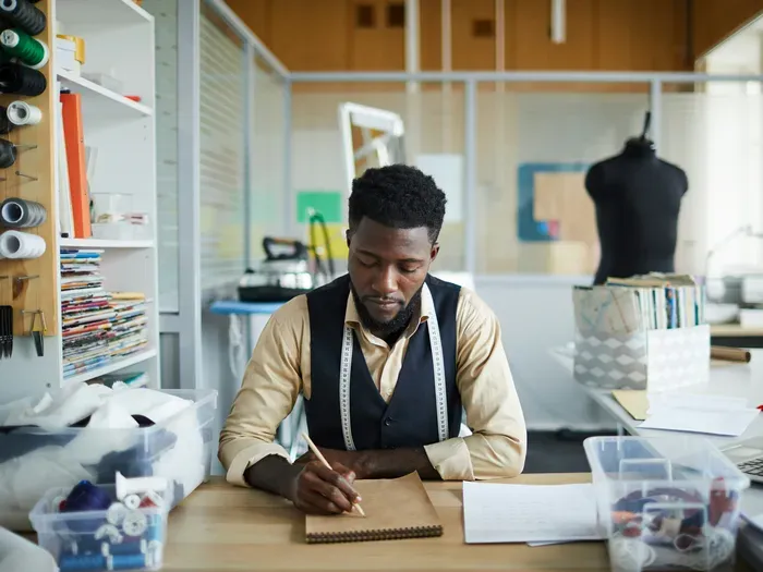 Male fashion designer sketching clothing ideas at his studio table surrounded by sewing tools, featured on towndirectorynow.com